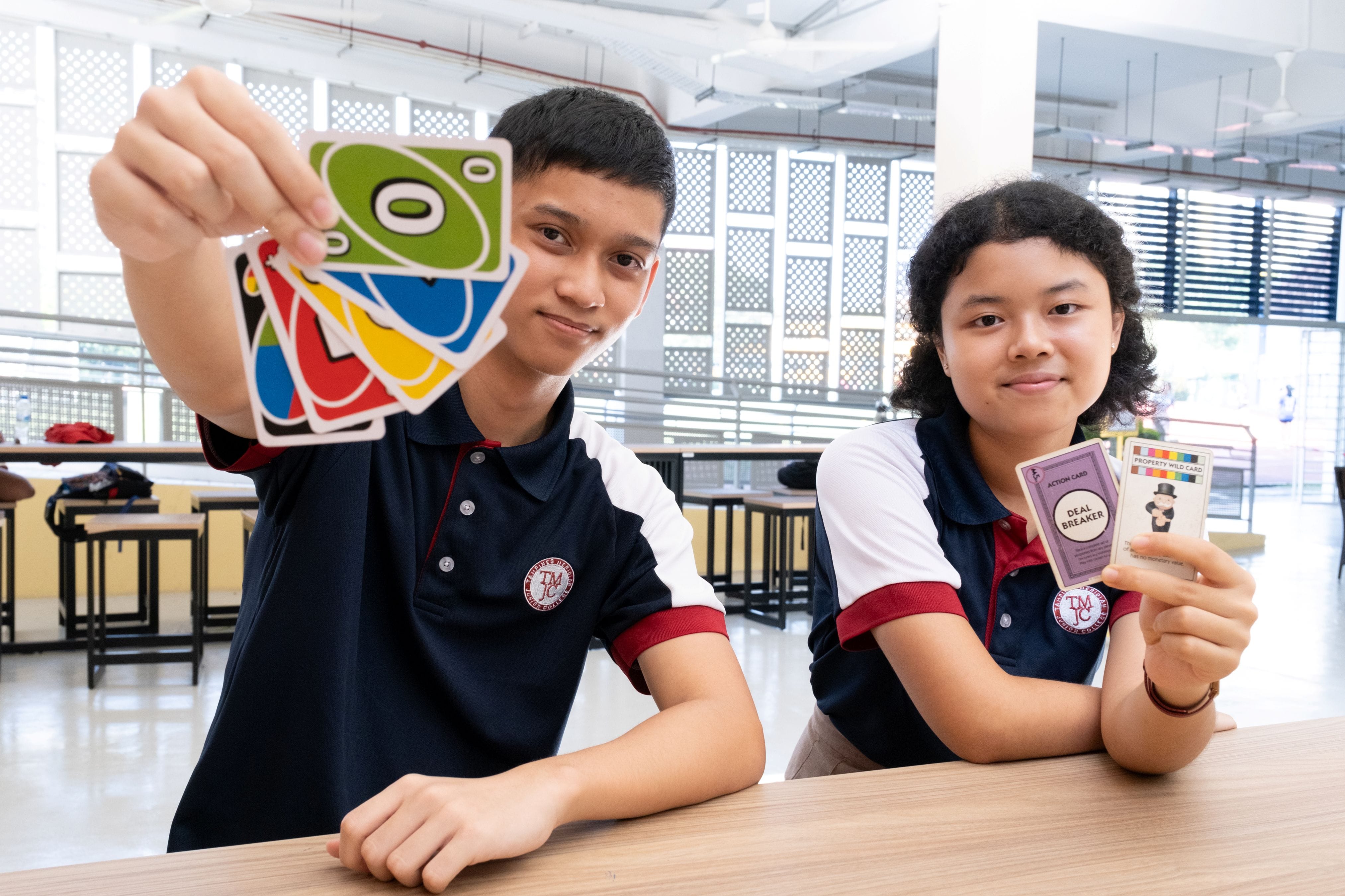 Two students in navy-and-white TMJC polo shirts sit at a table indoors. The student on the left holds up a fan of colourful UNO cards towards the camera, while the student on the right holds up two Monopoly Deal cards, including one labelled "Deal Breaker."